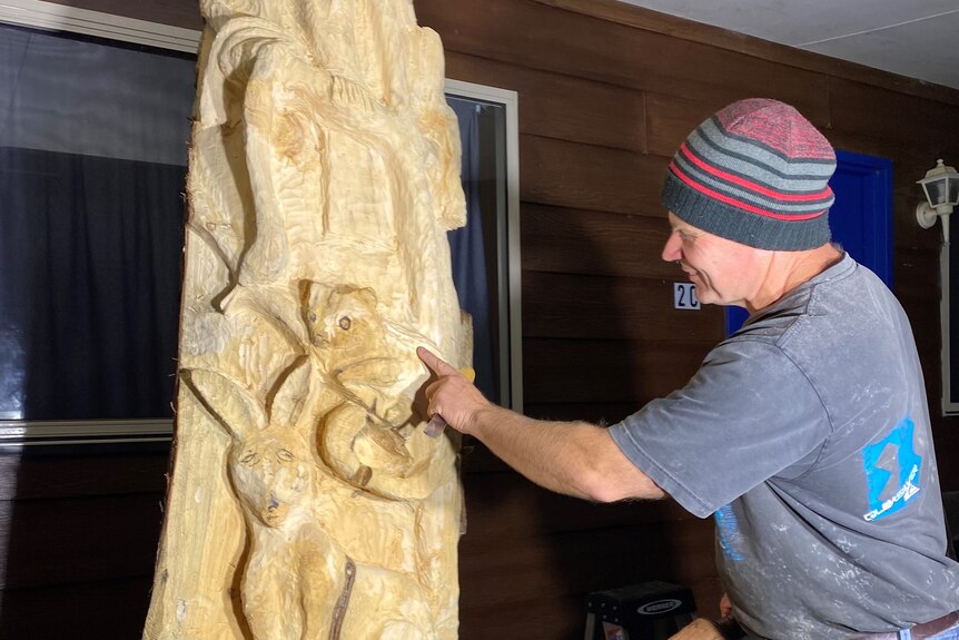 A man in a beanie and a grey t-shirt works works on a carved tree trunk. 