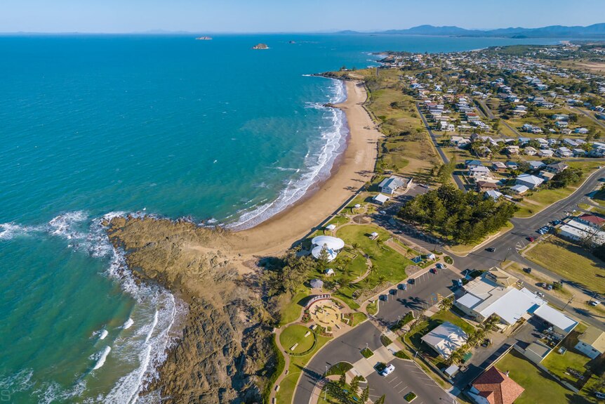 An aerial shot, ocean and headland to the left, houses, road and grass to the right.