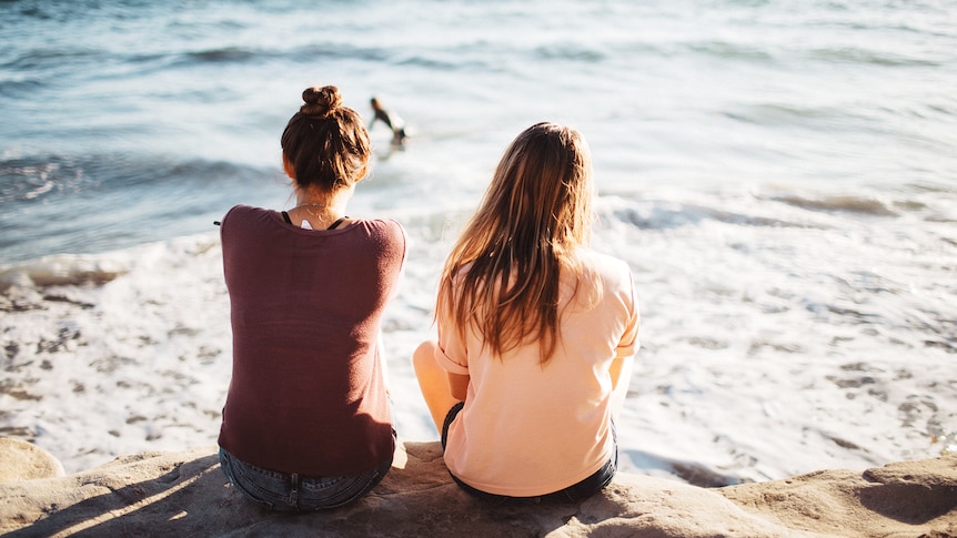 Two young women sitting on a rock by the ocean to depict how to make and keep friends after leaving high school.