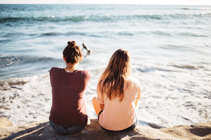 Two young women sitting on a rock with the ocean out of focus in the background.