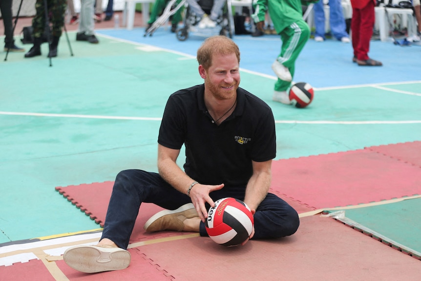 A close up of Prince Harry dressed in a black shirt and jeans sitting on the ground with a ball.