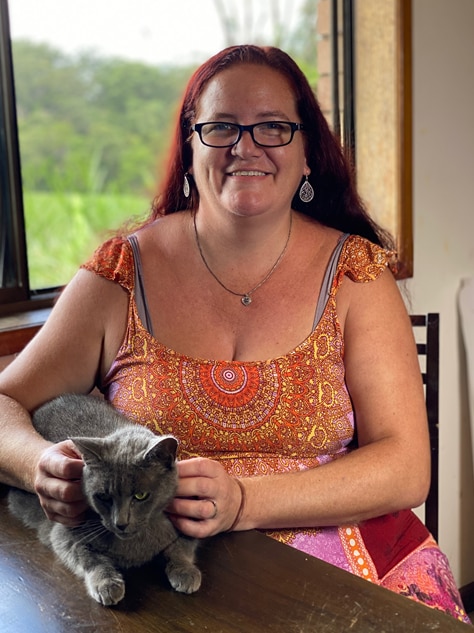 Middle aged woman with long red hair and big smile sitting at table, patting grey cat.