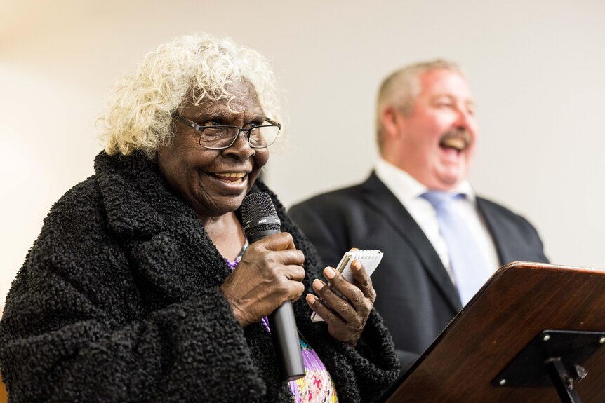 An elderly indigenous woman giving a speech as a man in a suit laughs at her joke.  