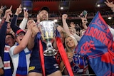 Max Gawn holds teh AFL premiership trophy and poses with fans holding a Melbourne Demons flag