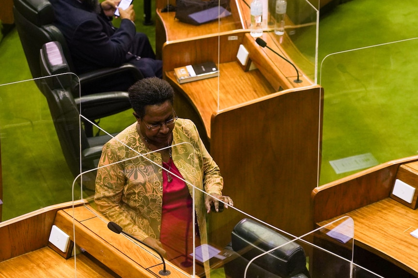 A woman wearing a floral printed coat over a pink dress walks into her seat in the green-carpeted house of parliament