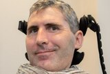 A smiling man with short grey hair, wearing a scarf and sitting in a wheelchair. An apparatus supports his neck.