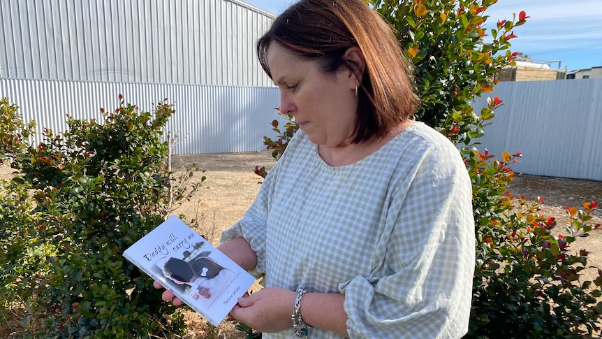 A woman holding a book looking down at it. There are trees and fences behind her.