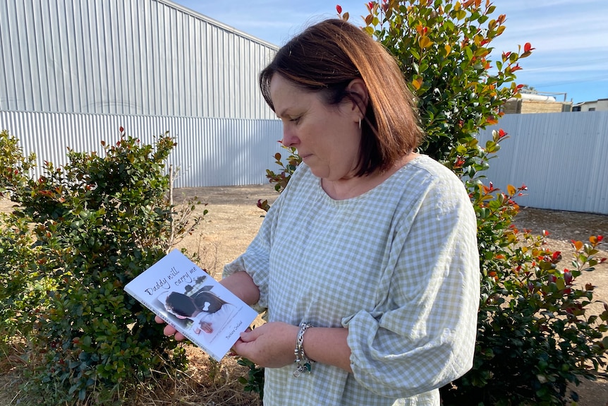 A woman holding a book looking down at it. There are trees and fences behind her.