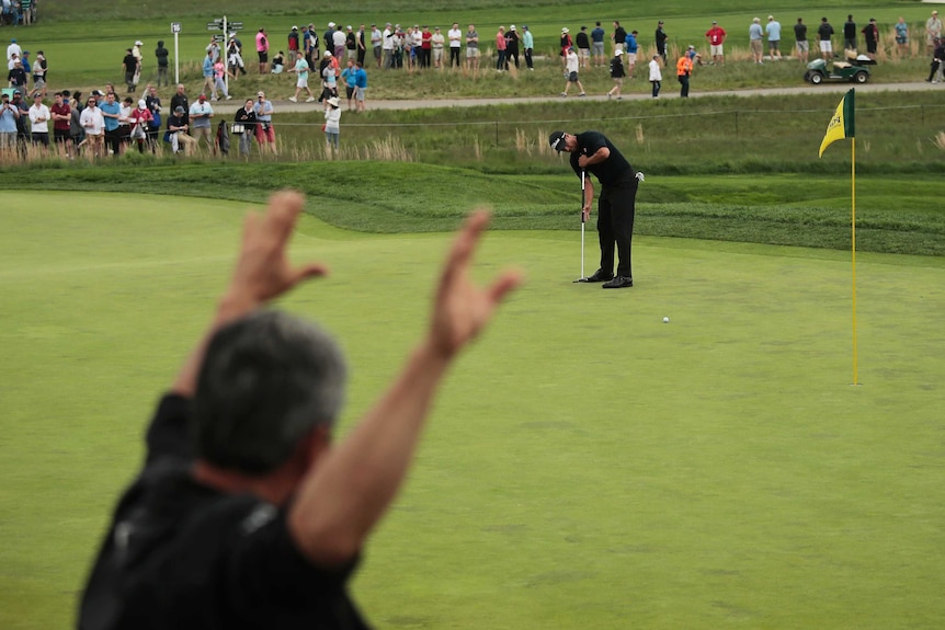 Golfer putts on the green, while an official holds his hands up to quieten the crowd.