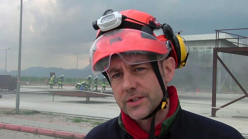 A man wearing a red helmet stands in front of a rescue drill being carried out in the background.