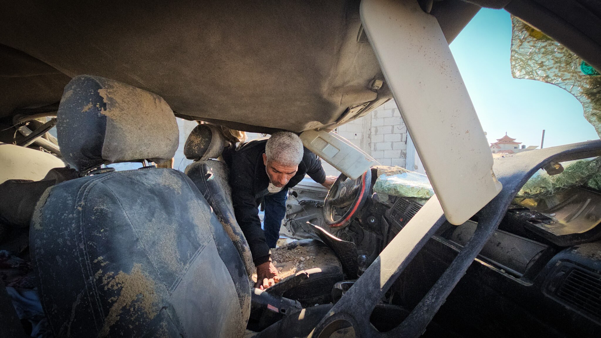 A man looks down while searching a destroyed car on a sunny day.
