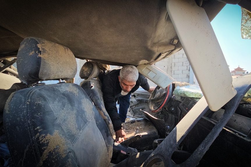 A man looks down while searching a destroyed car on a sunny day.