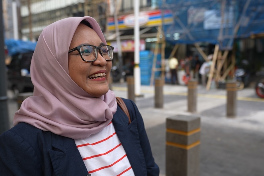 A woman wearing hijab and glasses smiling to the side when walking on a side walk.