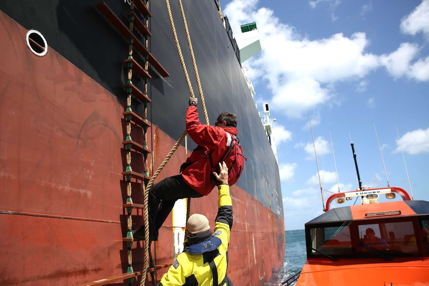 A man climbs up a rope ladder on the side of a container ship as another man watches on.