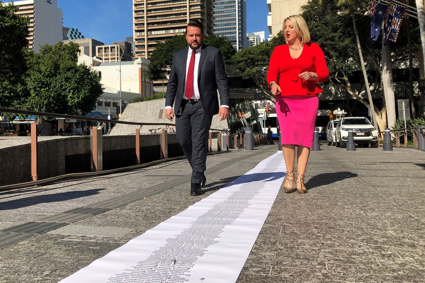 A man and a woman walk along King George Square looking at a paper rolled across the ground.