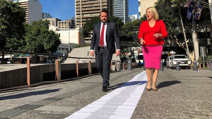 A man and a woman walk along King George Square looking at a paper rolled across the ground.