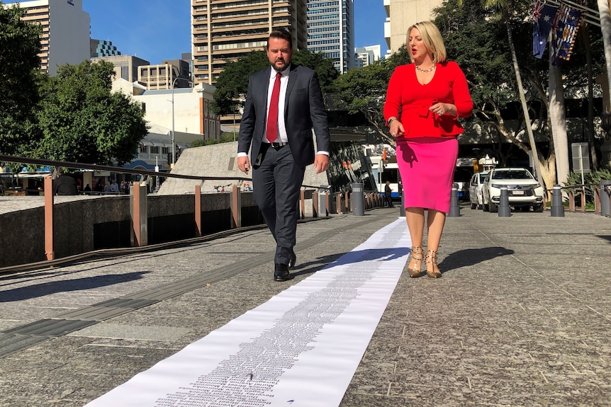 A man and a woman walk along King George Square looking at a paper rolled across the ground.