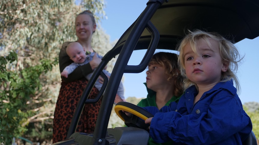 A little boy sits in a little car next to his brother while his mother stands in the background holding a baby