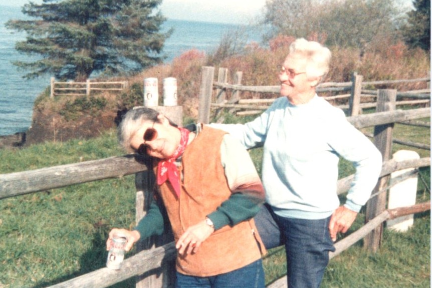 Two women smiling widely, standing near a wooden fence looking in different directions, with ocean in the distance behind them.