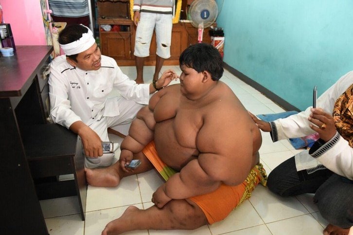 Arya Permana sits on the ground as a man squatting next to him takes a picture of him.