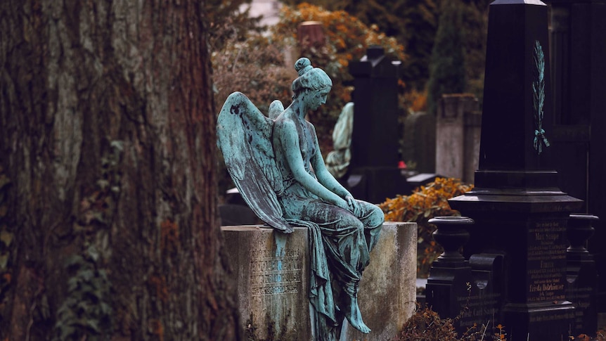 A statue of an angel appearing downcast in a cemetery surrounded by gravestones