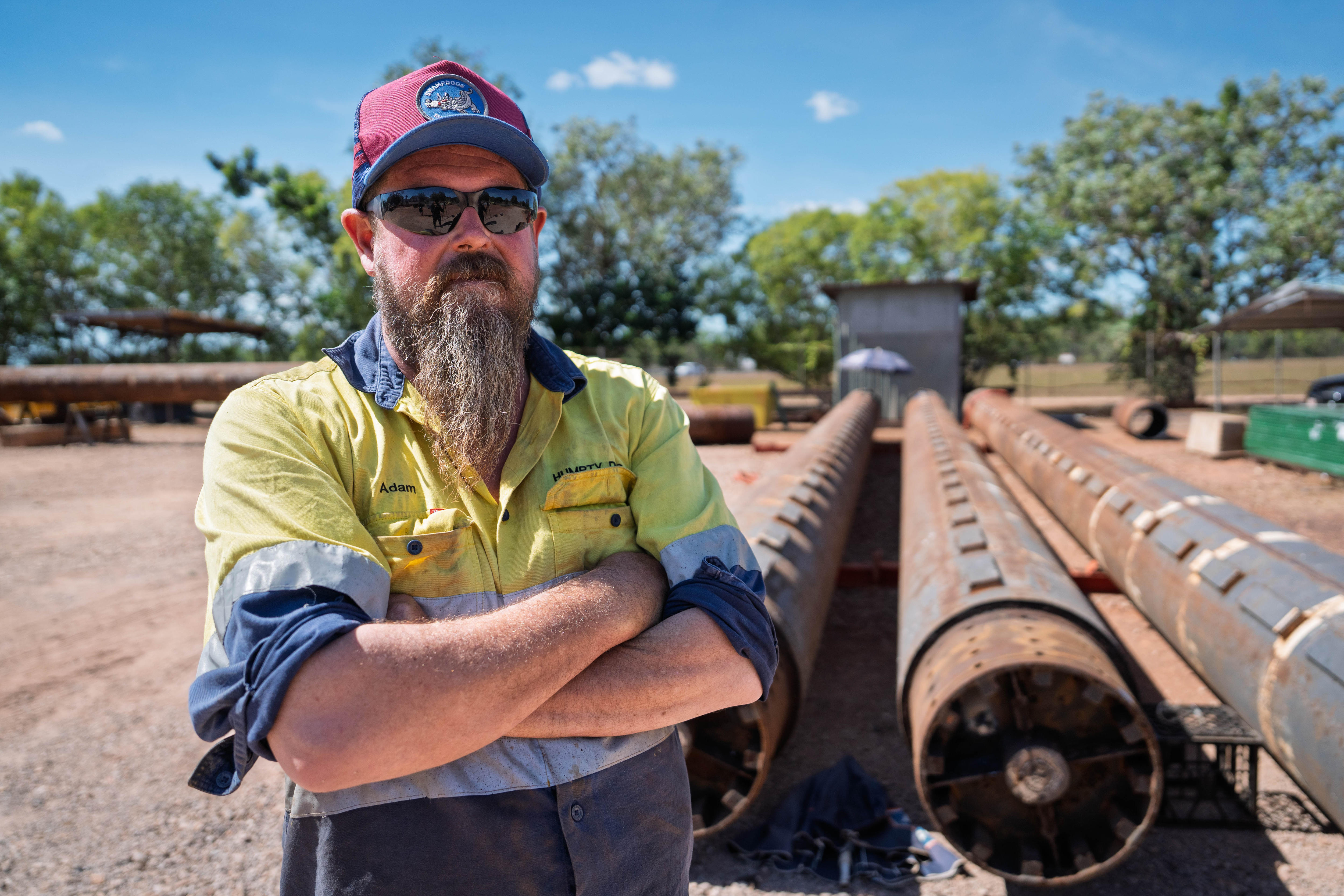 A man standing with his arms folded, in front of several long metal pipes in an outdoor work yard.