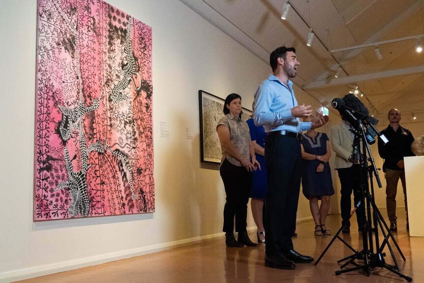 man in blue business shirt stands in front of microphones inside art gallery
