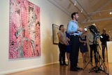 A man in blue business shirt stands in front of microphones inside art gallery.
