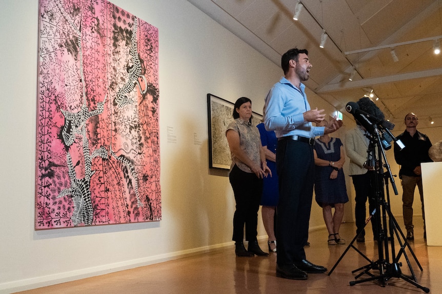 man in blue business shirt stands in front of microphones inside art gallery