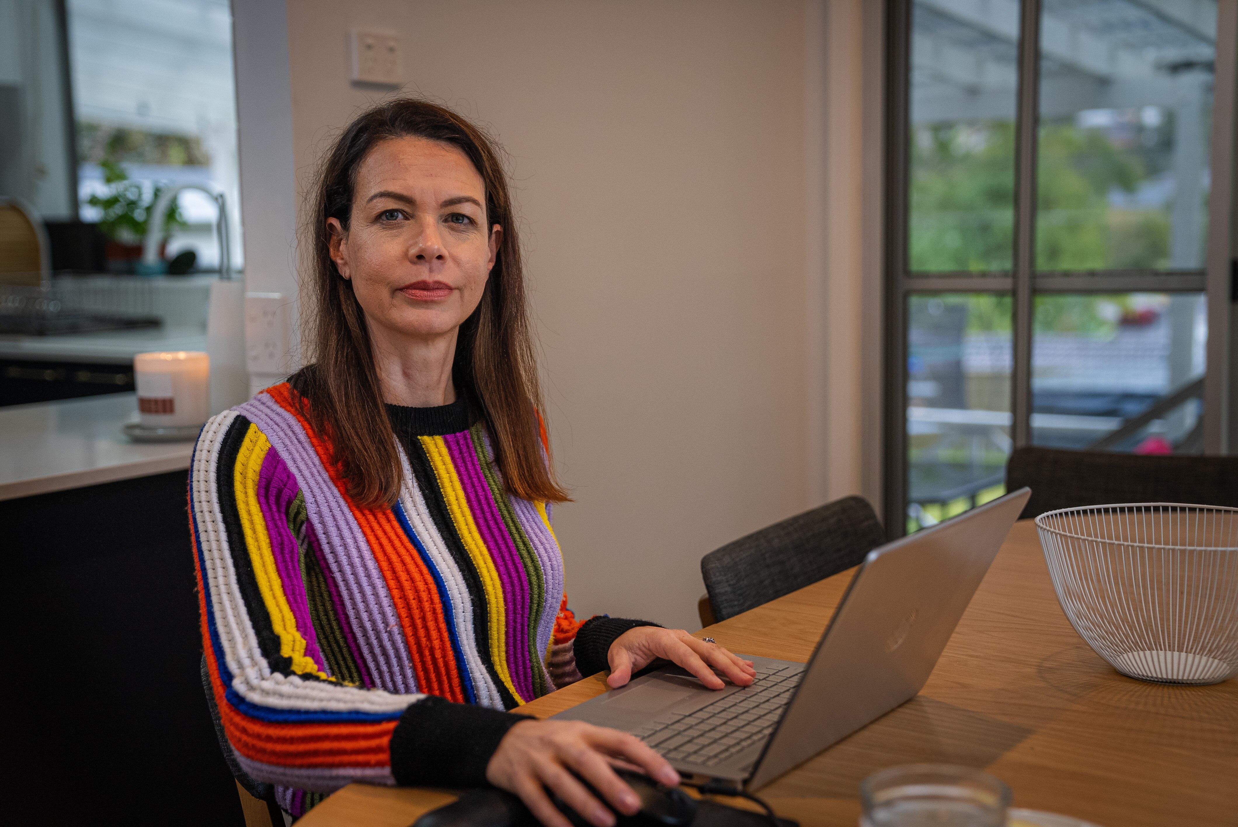 A woman with dark hair, wearing a rainbow-coloured top, working on her laptop.