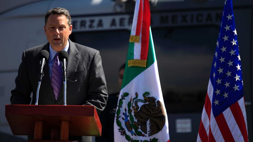 John Feeley stands in front of an American and Mexican flag and delivers a speech.