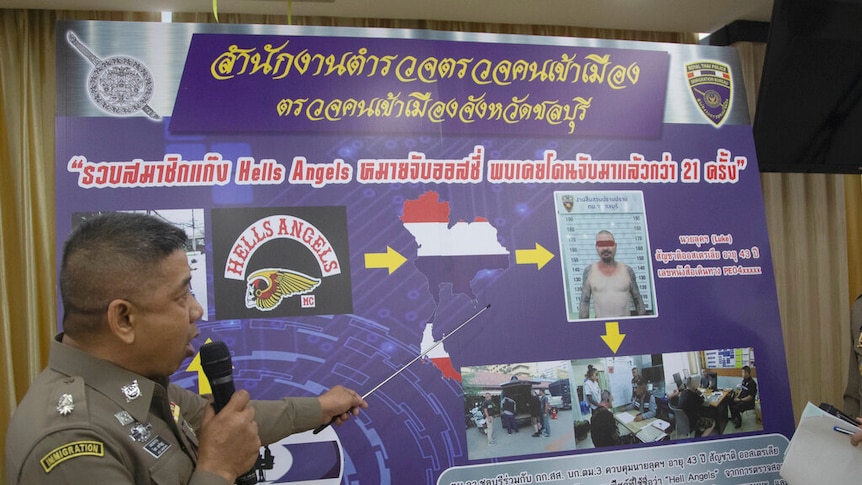 A Thai immigration officer holds a microphone and pointer in front of a large information board detailing criminal information.