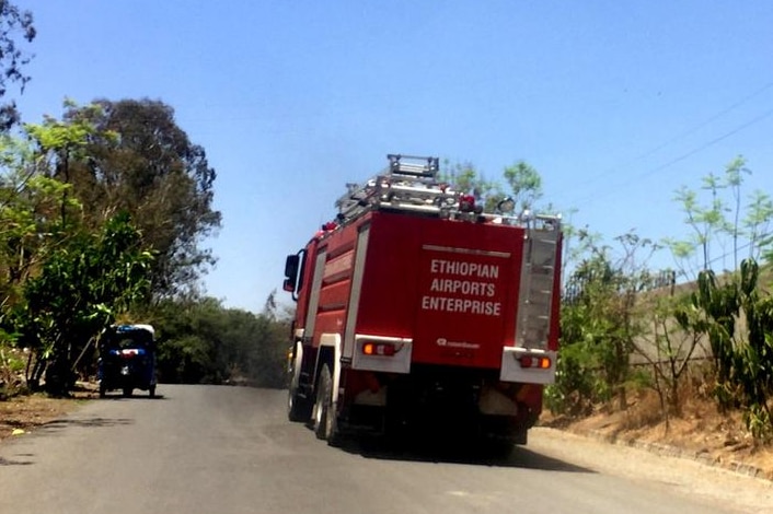 Red firetruck drives on road with trees lining it. Skies are blue overhead and there is a tuk tuk on the left hand side of road.