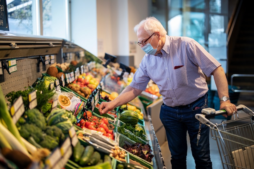 A senior man with short white hair and a face mask browses the fruit and vegetable at the grocery store