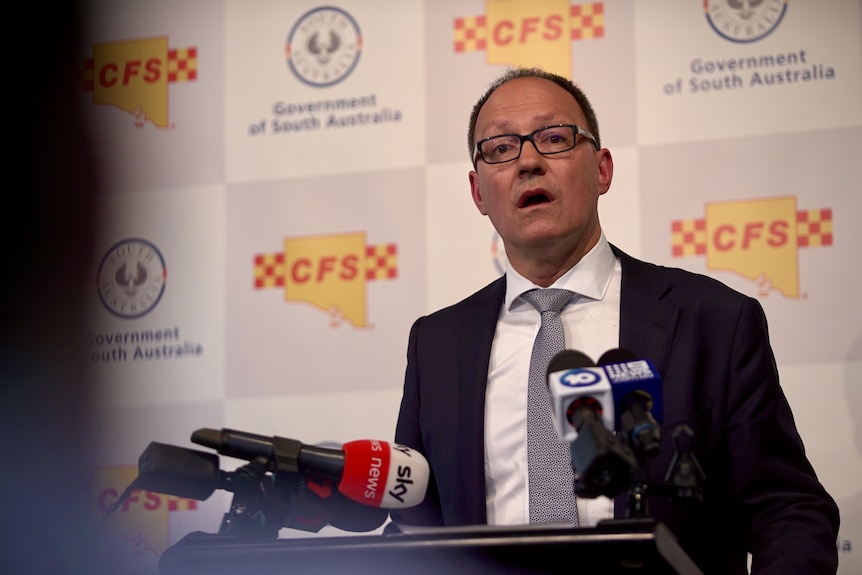 A man with glasses in a suit looking into a crowd at a press conference with his mouth open