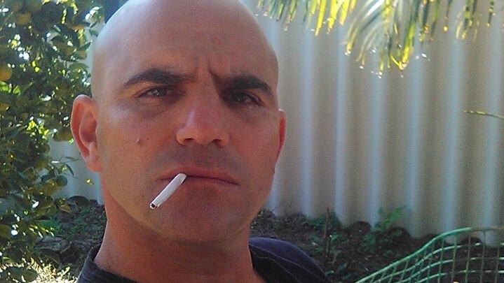 A selfie of Milos Radovic with a cigarette hanging out of his mouth wearing a blue shirt outdoors.