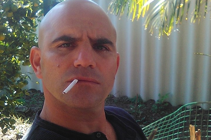 A selfie of Milos Radovic with a cigarette hanging out of his mouth wearing a blue shirt outdoors.