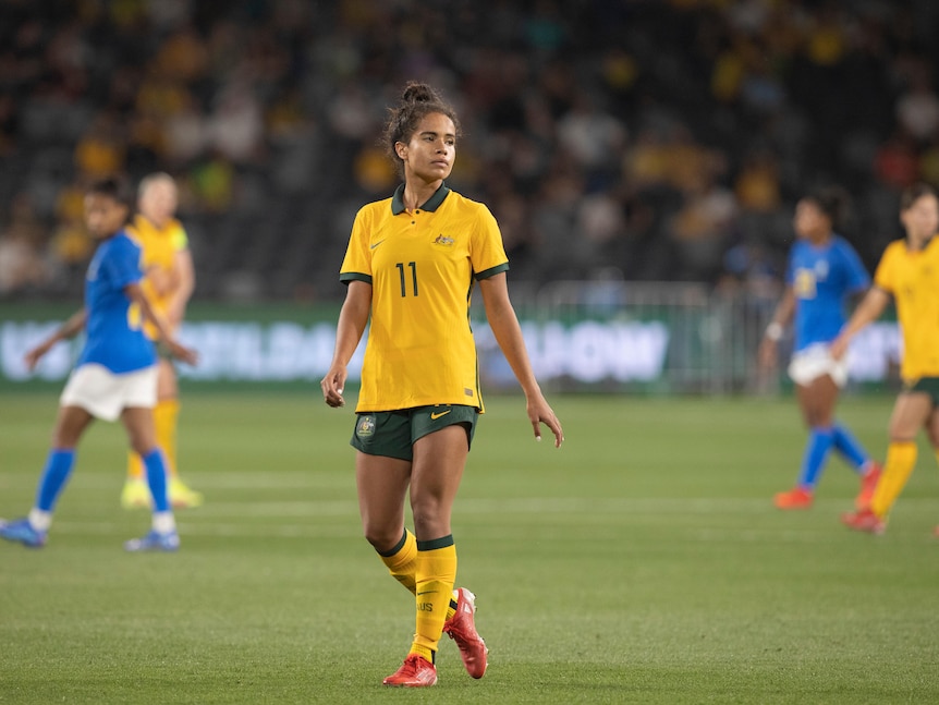 A female soccer player wearing yellow and green looks across the field during a game
