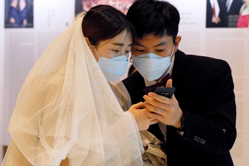 A formally dressed couple, wearing face masks, check their shared mobile telephone in a hall.