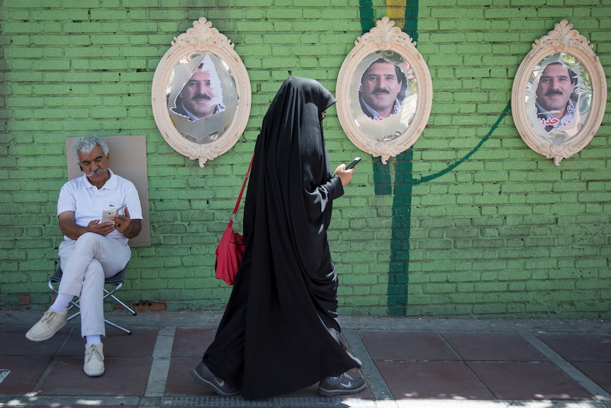 A woman in a full hijab and robe walks past a man sitting down outside, while looking at her phone
