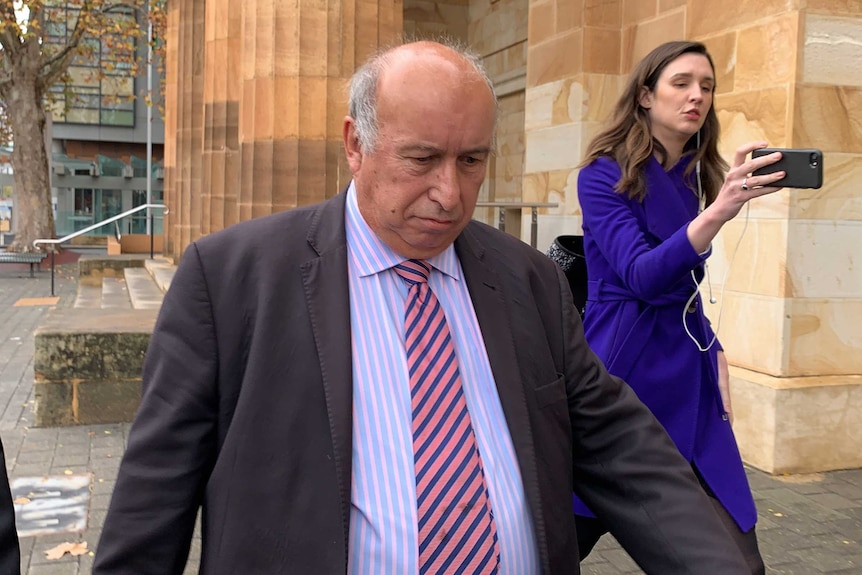 A man wearing a suit walks in front of a sandstone building while a woman takes his photo on a mobile phone