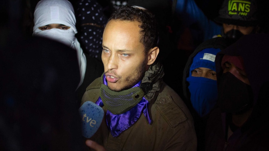 Oscar Perez speaks to the press at a night vigil in Caracas, Venezuela. The people behind him are wearing masks, he is not.