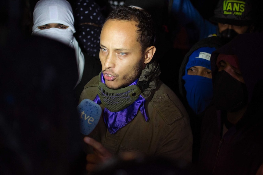 Oscar Perez speaks to the press at a night vigil in Caracas, Venezuela. The people behind him are wearing masks, he is not.