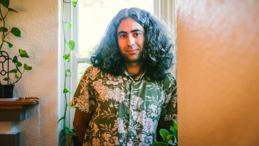 A man with shoulder-length hair and a green shirt standing in a hallway of a house.