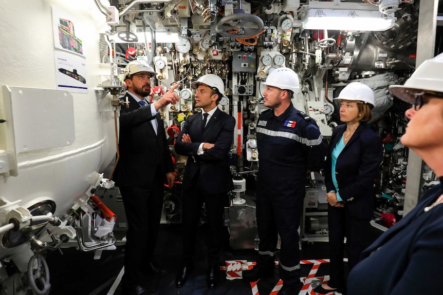 Emmanuel Macron folds his arms as a guide shows him around a nuclear submarine as onlookers look on