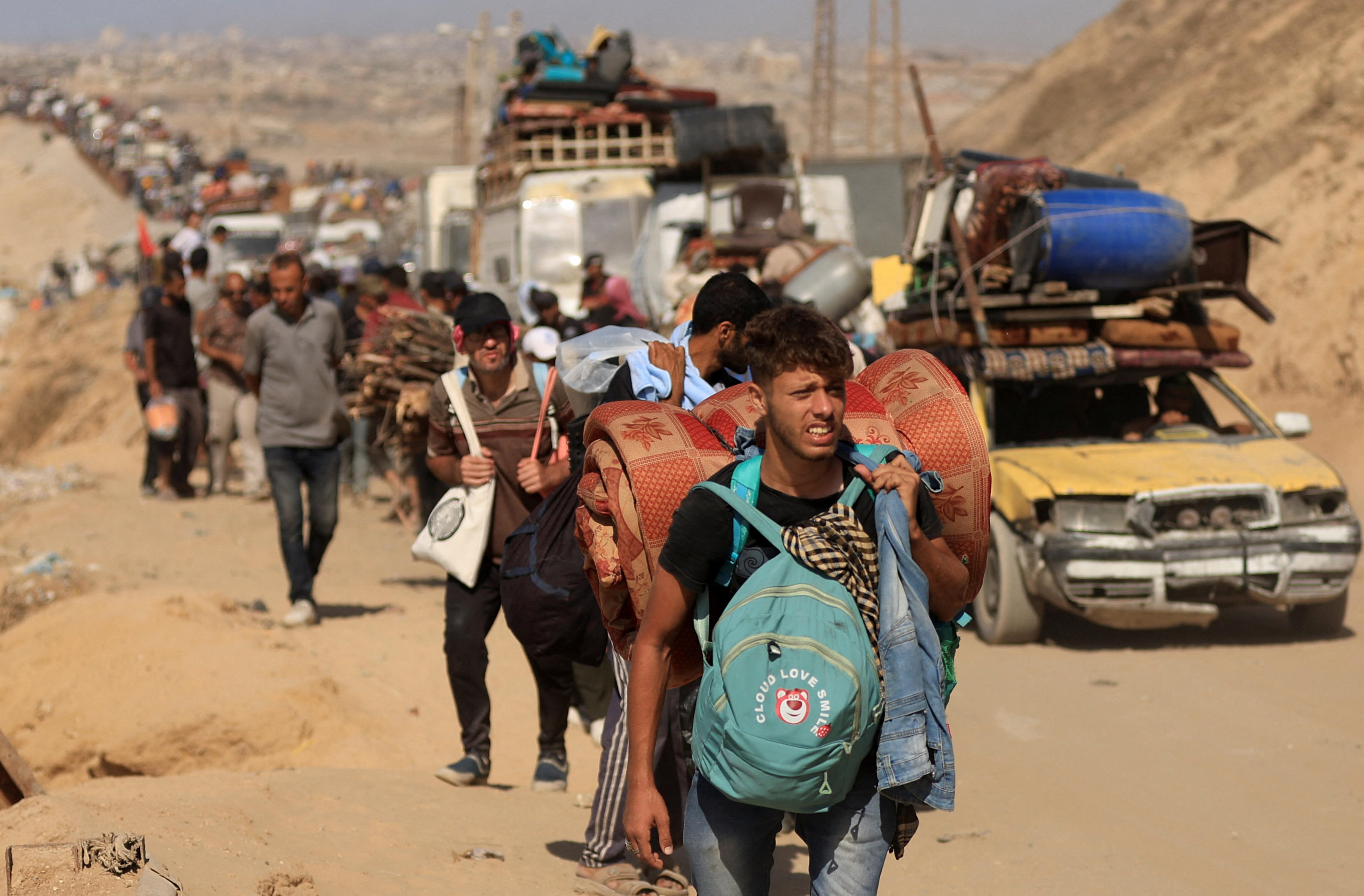 A long line of people walk down a dusty road carrying bags.