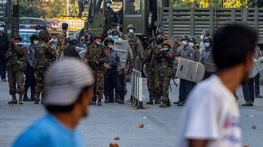 A line of soldiers, one pointing his gun in the direction of the camera. In the foreground stand two men, out of focus. 