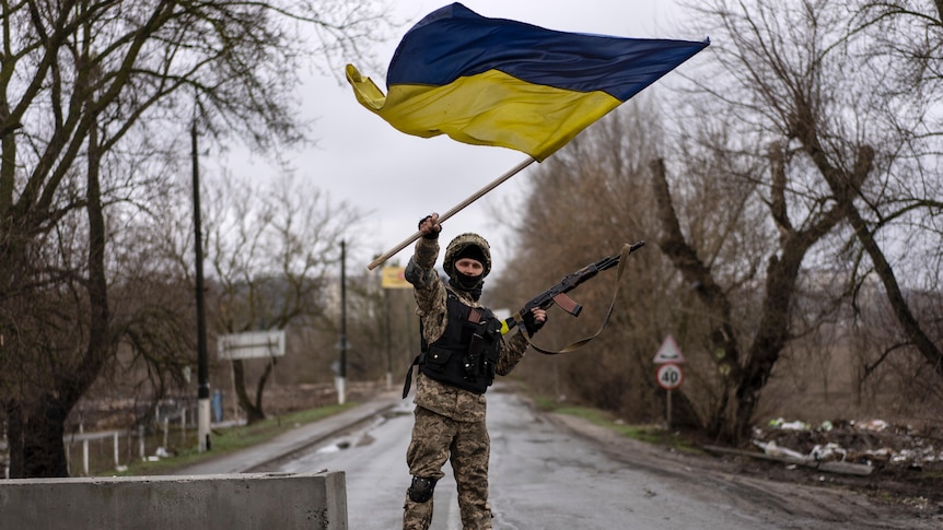 一名士兵站在混凝土块上，左手拿着步枪，右手挥舞着乌克兰国旗。