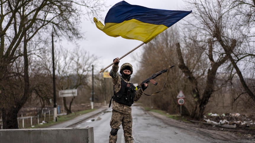 A soldier waves a Ukrainian flag with his right hand as he holds a rifle in his left hand while standing atop a concrete block.