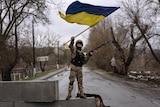 An soldier waves a Ukrainian flag with his right hand as he holds a rifle in his left hand while standing atop a concrete block.